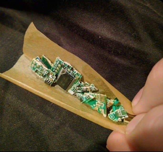 A hand holds a brown paper cone containing a pile of small, green circuit boards and electronic components, resembling a takeout food container. The image is a photograph with no visible text.