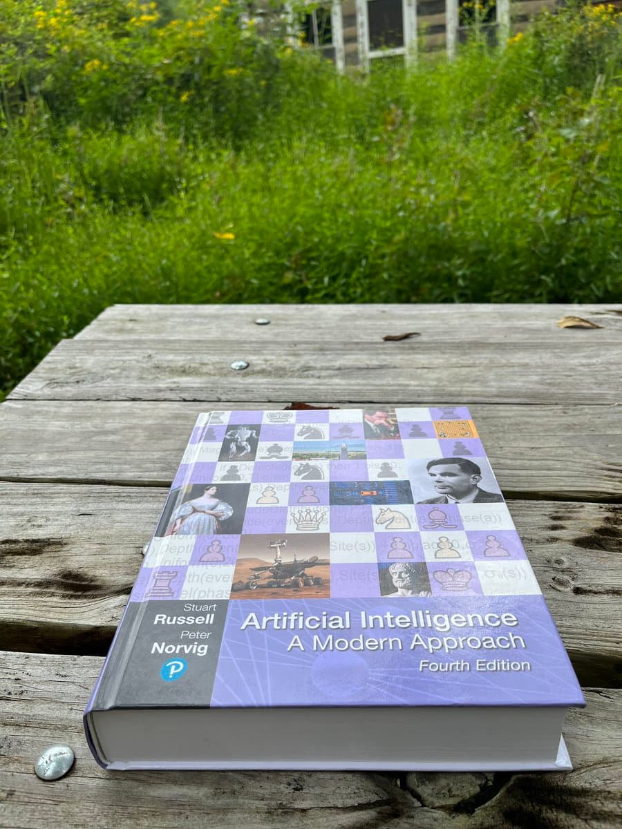 A photograph of the book "Artificial Intelligence: A Modern Approach, Fourth Edition" by Stuart Russell and Peter Norvig, lying on a weathered wooden table outdoors with green foliage in the background.