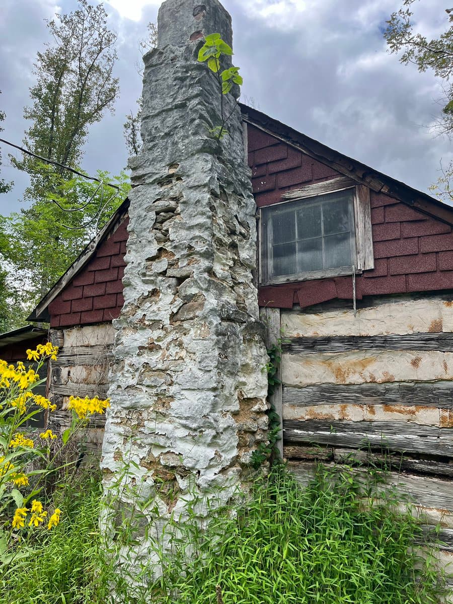 A weathered, crumbling stone chimney stands in front of an old log cabin with red shingles and a small window. The structure is surrounded by overgrown green grass and yellow wildflowers under a cloudy sky.