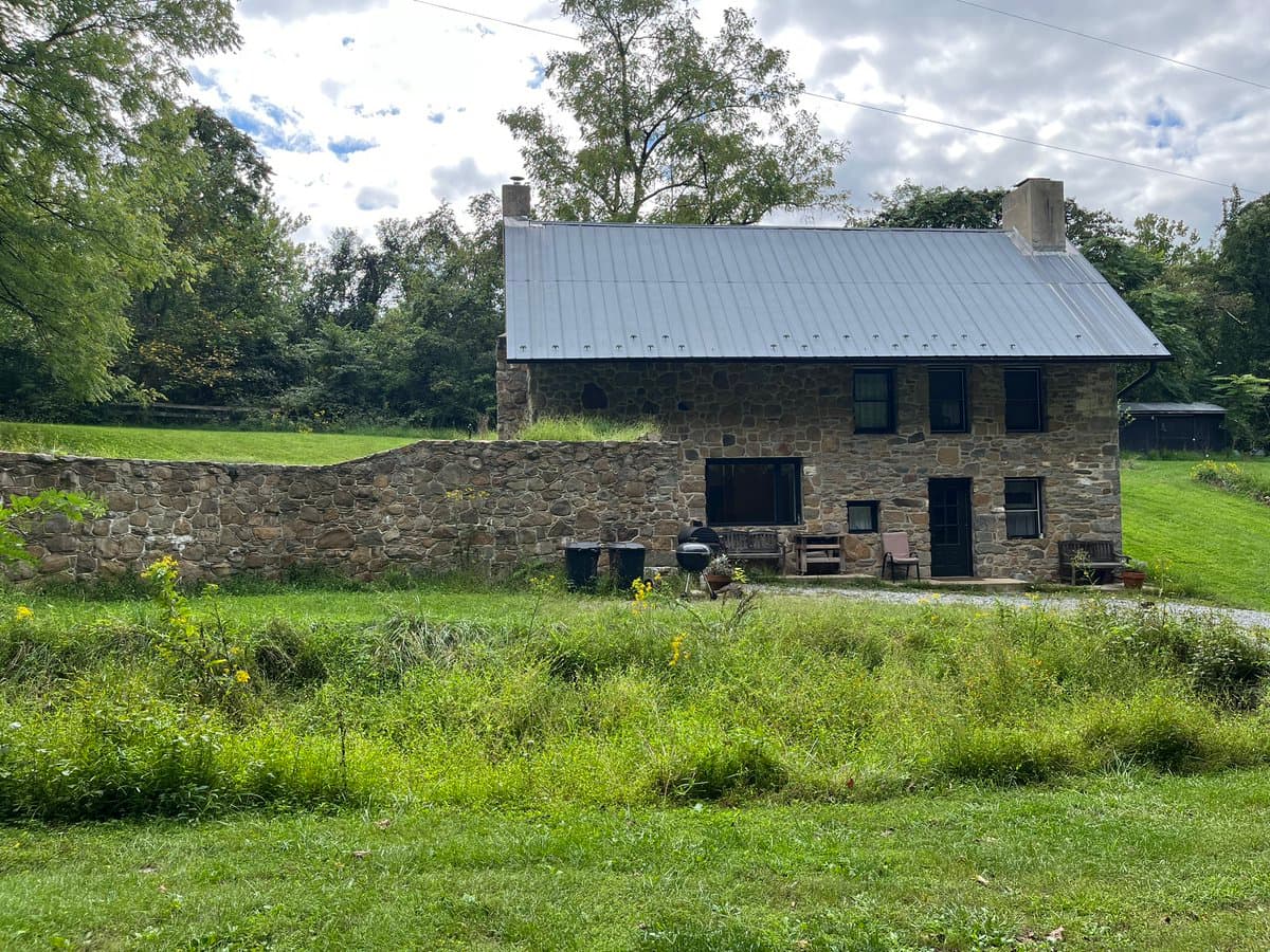 A photograph of a two-story stone house with a metal roof and a matching stone wall, set in a grassy yard with trees in the background.