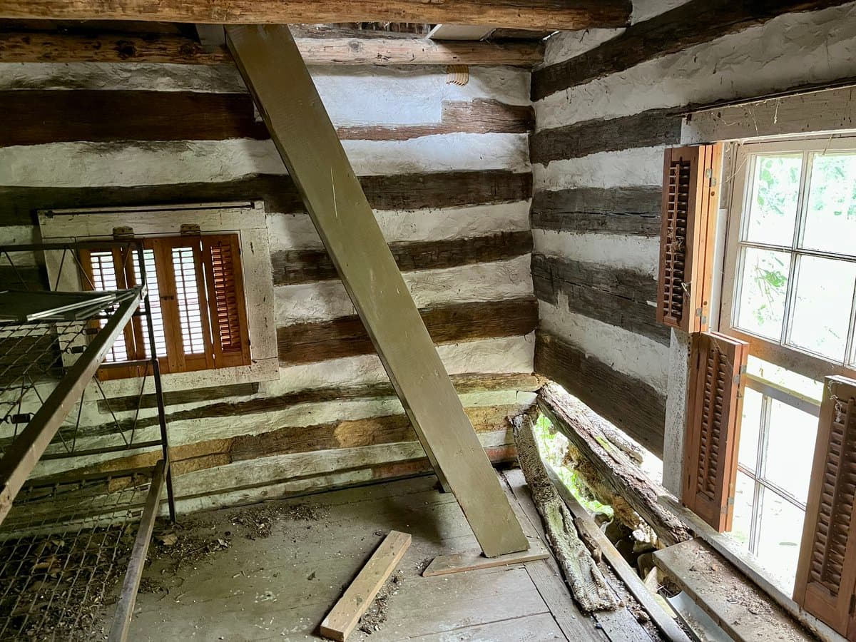 An interior photograph of a rustic attic space featuring log-and-chink walls, a sloped wooden ceiling, and a diagonal support beam. The room includes a wire-mesh loft structure on the left and two windows with wooden shutters on the right, one of which is open.