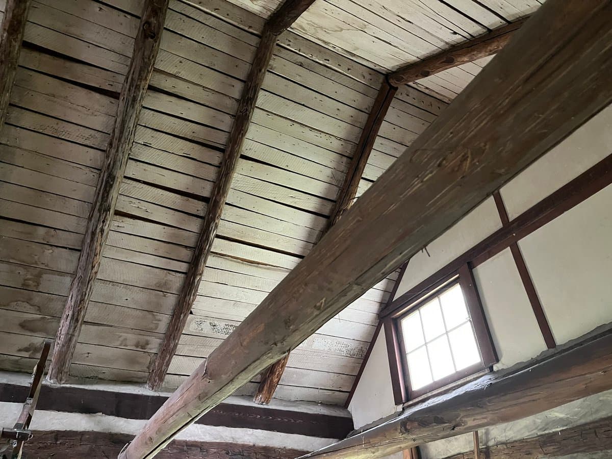 A low-angle interior photograph of a rustic wooden structure, showing a sloped ceiling made of weathered planks supported by rough-hewn timber beams. A small, multi-paned window is set into a white plaster wall on the right side.