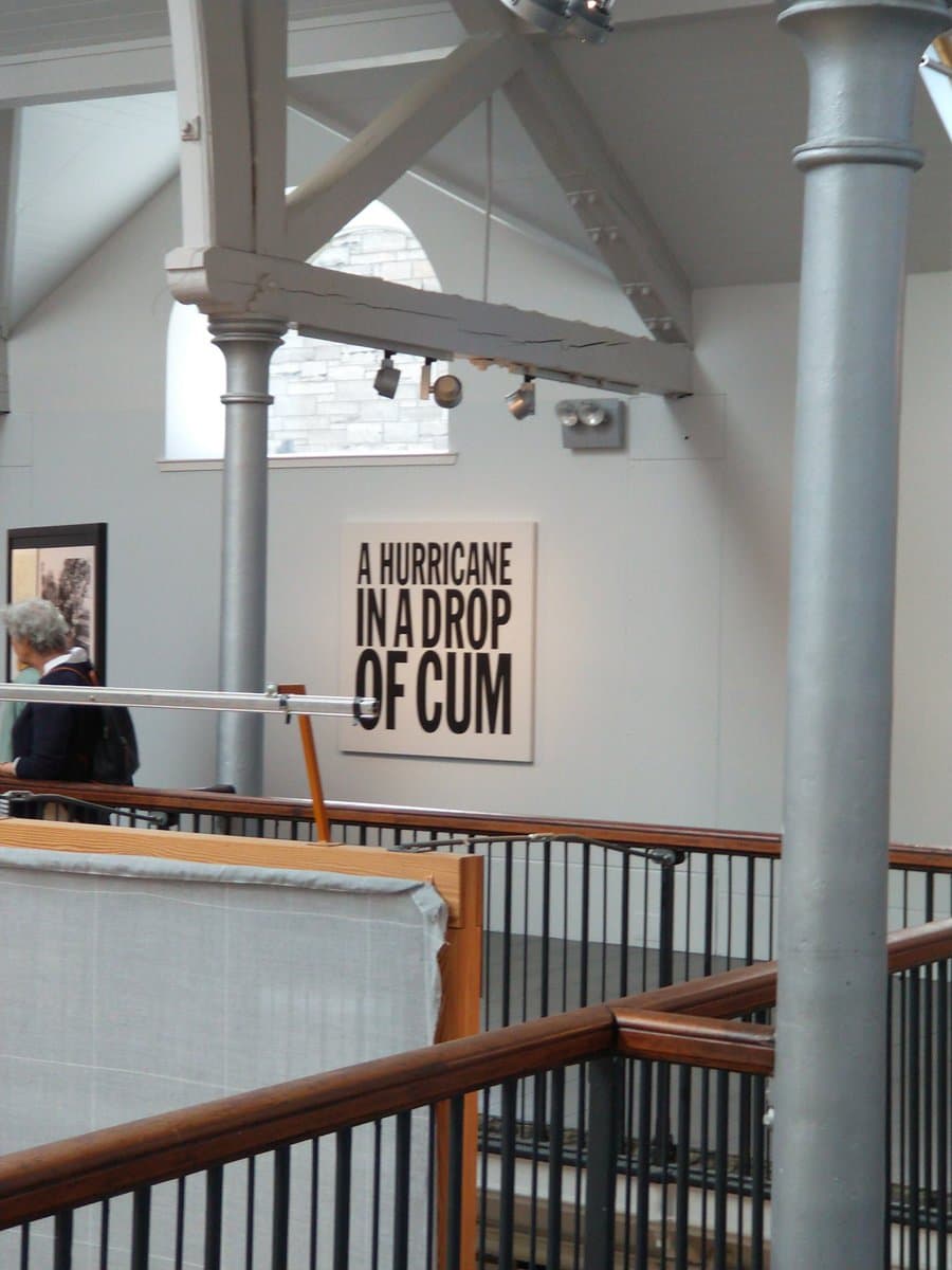 A photograph of an indoor gallery or museum space featuring a white wall with a large typographic poster. The room has high ceilings with exposed wooden beams, metal support columns, and a wooden railing in the foreground. A person is partially visible on the left side of the frame.