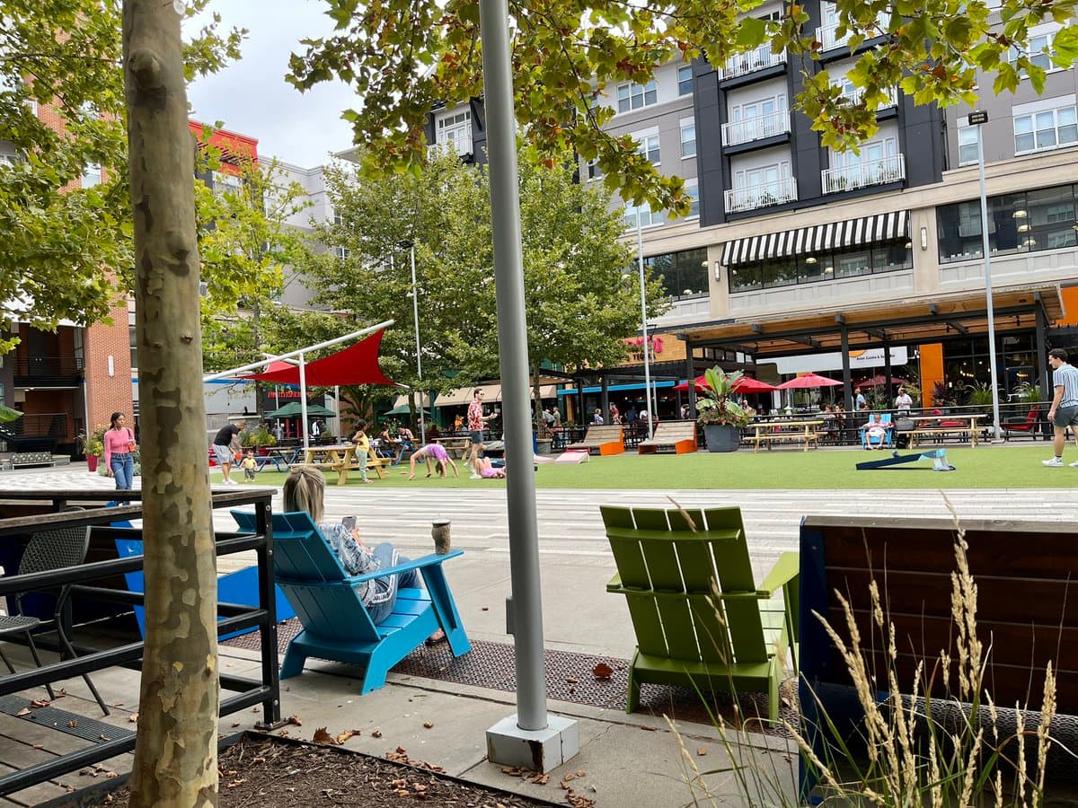 A wide-angle outdoor photograph of a public urban plaza featuring a large artificial grass lawn, colorful Adirondack chairs, and a red shade sail. People are relaxing on the lawn and sitting in the chairs, with a multi-story building and storefronts visible in the background.