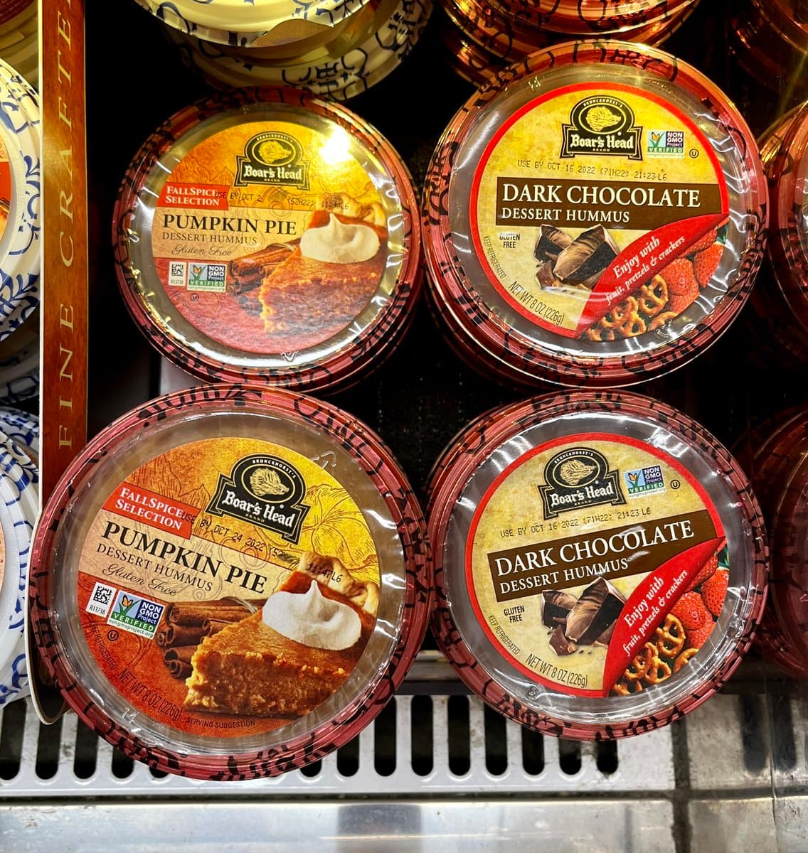 A close-up shot of four round containers of Boar's Head dessert hummus on a store shelf, featuring two varieties: Pumpkin Pie and Dark Chocolate. The labels display the product names, flavor descriptions, and serving suggestions.