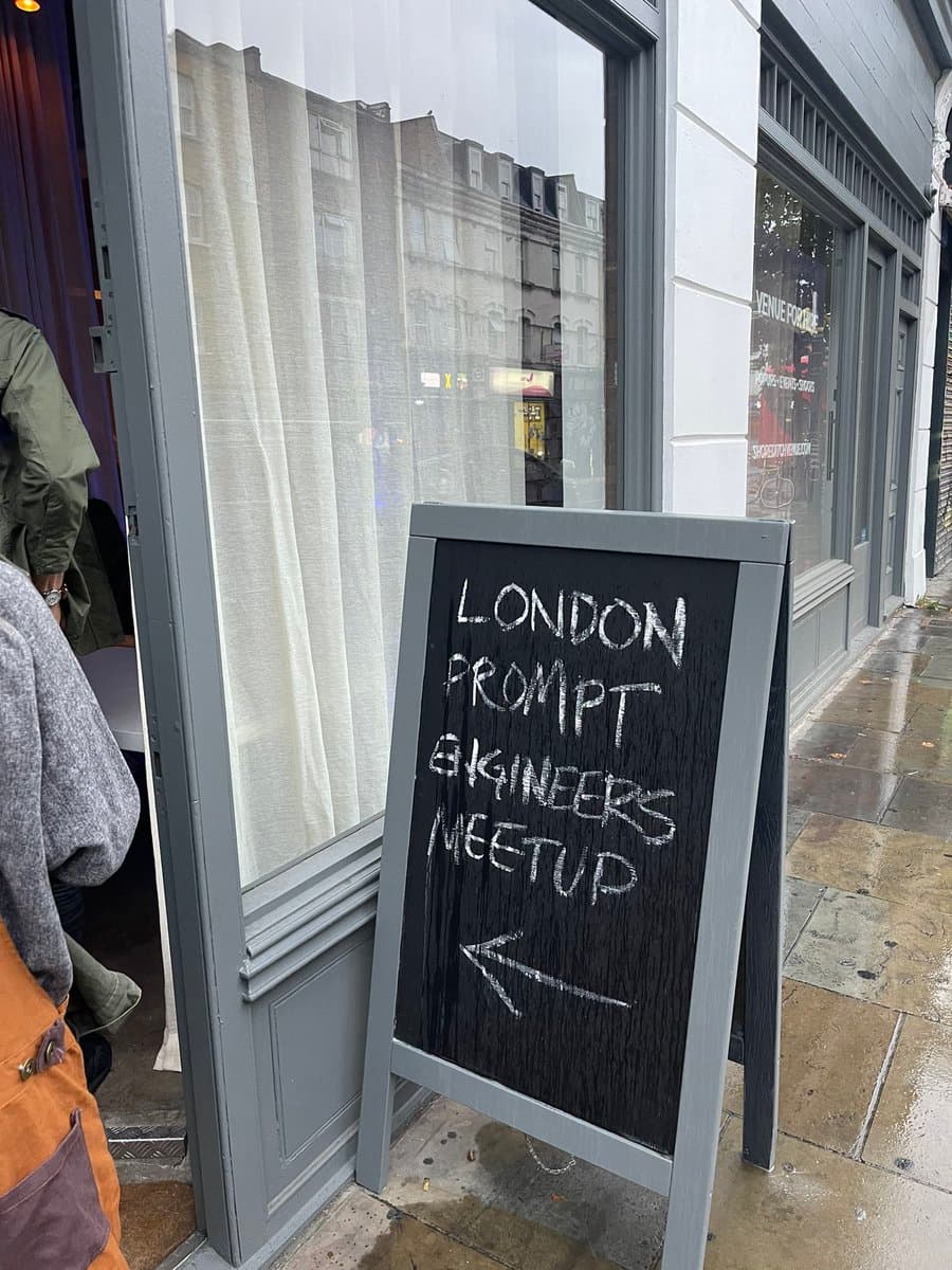 A black A-frame chalkboard sign stands on a wet sidewalk outside a storefront, advertising a local event. The sign is written in white chalk and points towards the entrance of the building.
