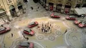 A high-angle, wide shot of a busy city plaza with several cars parked in a circular pattern around a central gathering of people. The scene appears to be in a European city, featuring ornate architecture and cobblestone streets.
