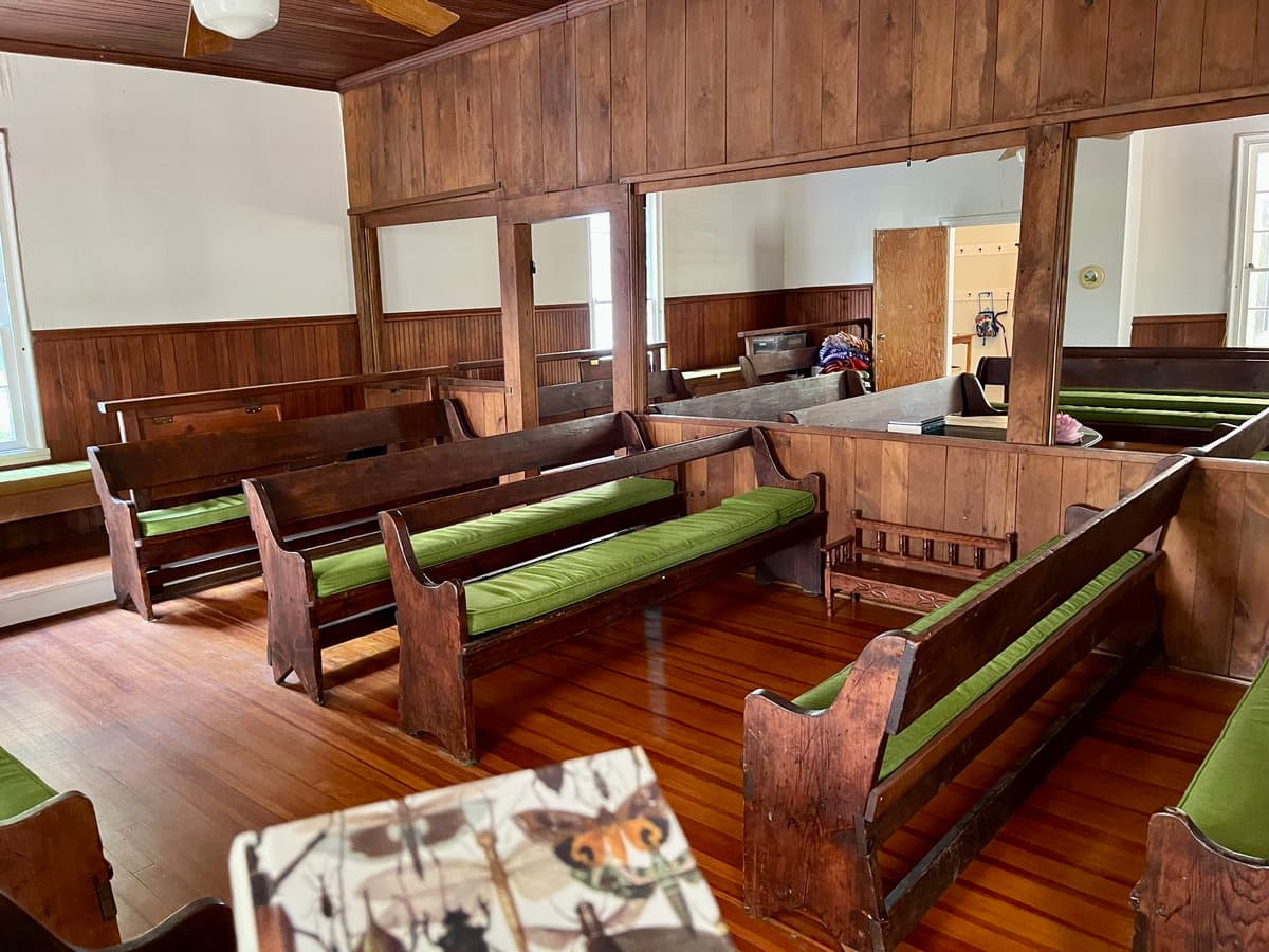 An interior photograph of a room with wooden paneling and a hardwood floor, furnished with several rows of wooden benches featuring green cushions. The room has large open window frames and a ceiling fan, with a butterfly-themed book or pamphlet visible in the foreground.