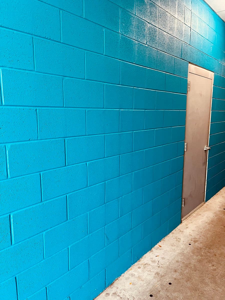A photo of a cinder block wall painted a bright turquoise blue, with a plain brown door set into the right side of the wall. The floor is bare concrete.