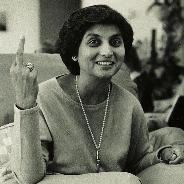 A black-and-white photograph of a smiling woman with short dark hair, wearing a long beaded necklace and a ring, raising her index finger while seated on a couch.