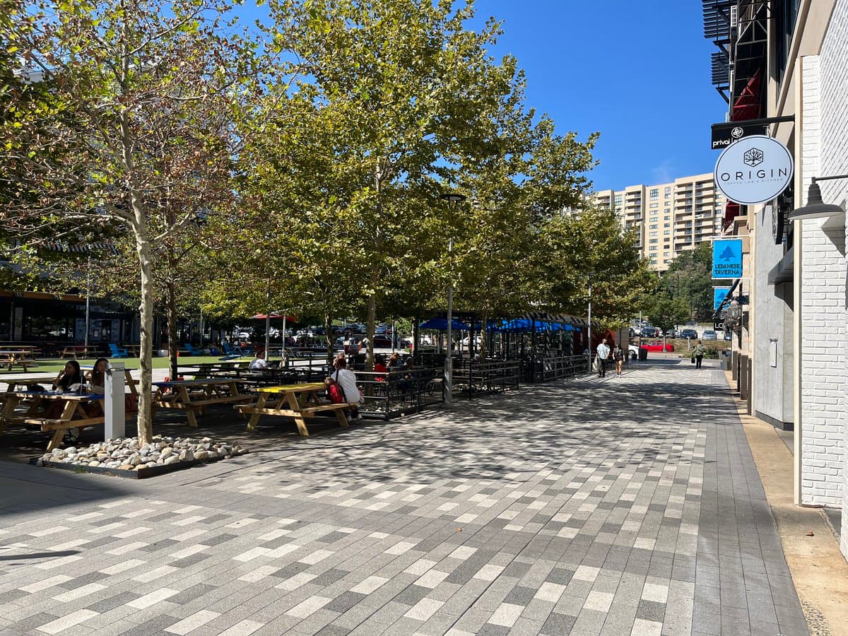A sunny outdoor plaza featuring a wide, patterned brick walkway lined with young trees and wooden picnic tables where several people are seated. In the background, a large multi-story residential building is visible, while a white brick building with a circular 'ORIGIN' sign stands on the right.