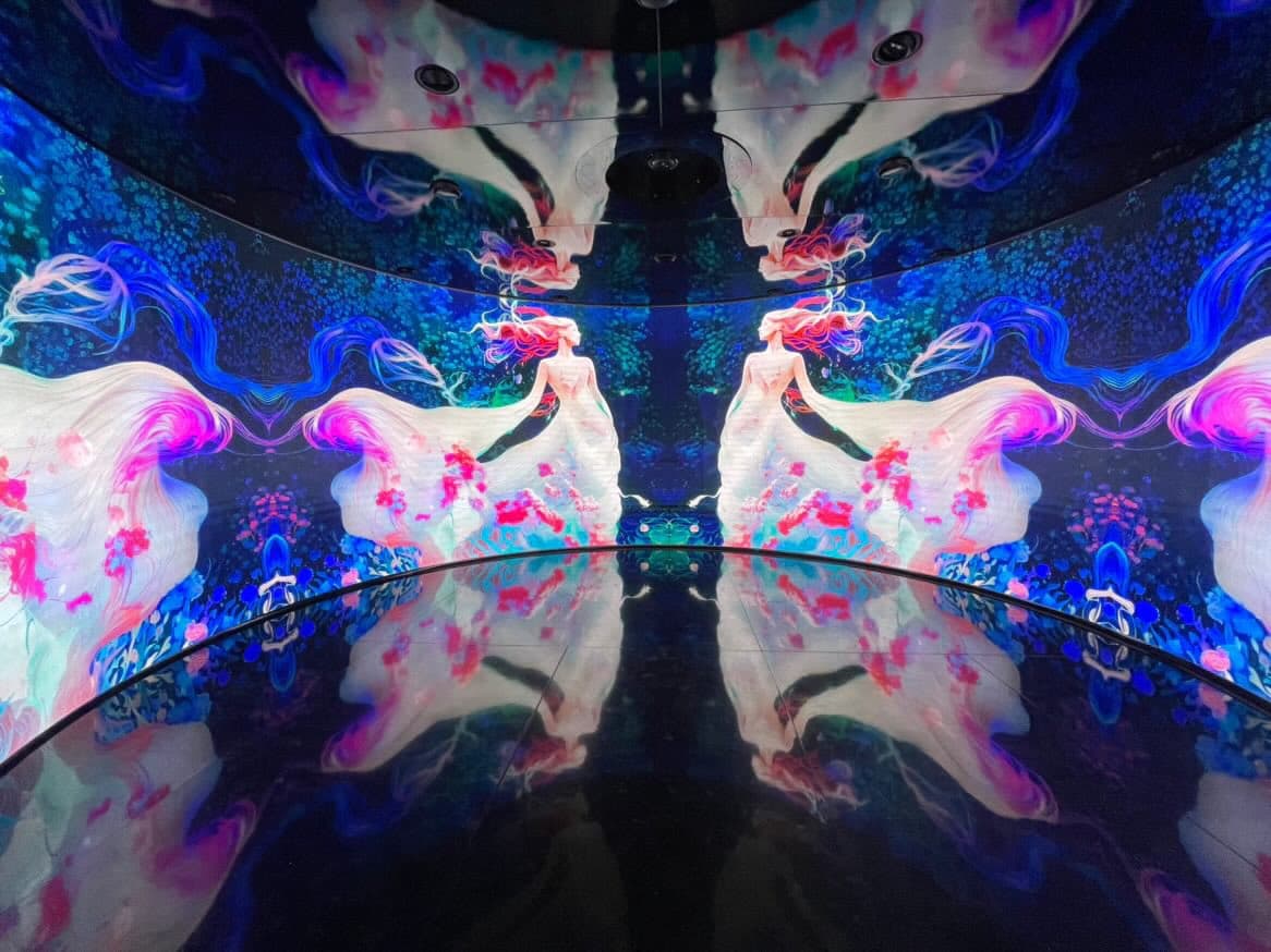 A vibrant, immersive digital art installation featuring a symmetrical, kaleidoscopic projection of a figure with flowing, colorful hair and garments. The artwork is displayed on curved walls and a reflective floor, creating a mirrored, infinite effect in a dark room.