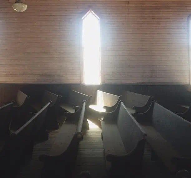A dimly lit interior of a wooden chapel or church, featuring rows of empty wooden pews and a tall, arched window that lets in bright light. The scene has a quiet, contemplative atmosphere.