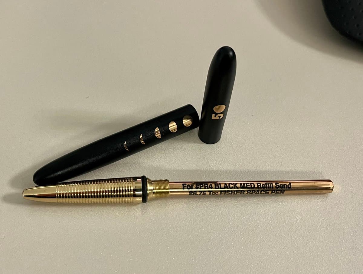 A close-up photograph of a disassembled black and gold Fisher Space Pen refill lying on a white surface. The image shows the gold refill cartridge, a black textured grip section, and a black cap with the number 50 printed in gold.