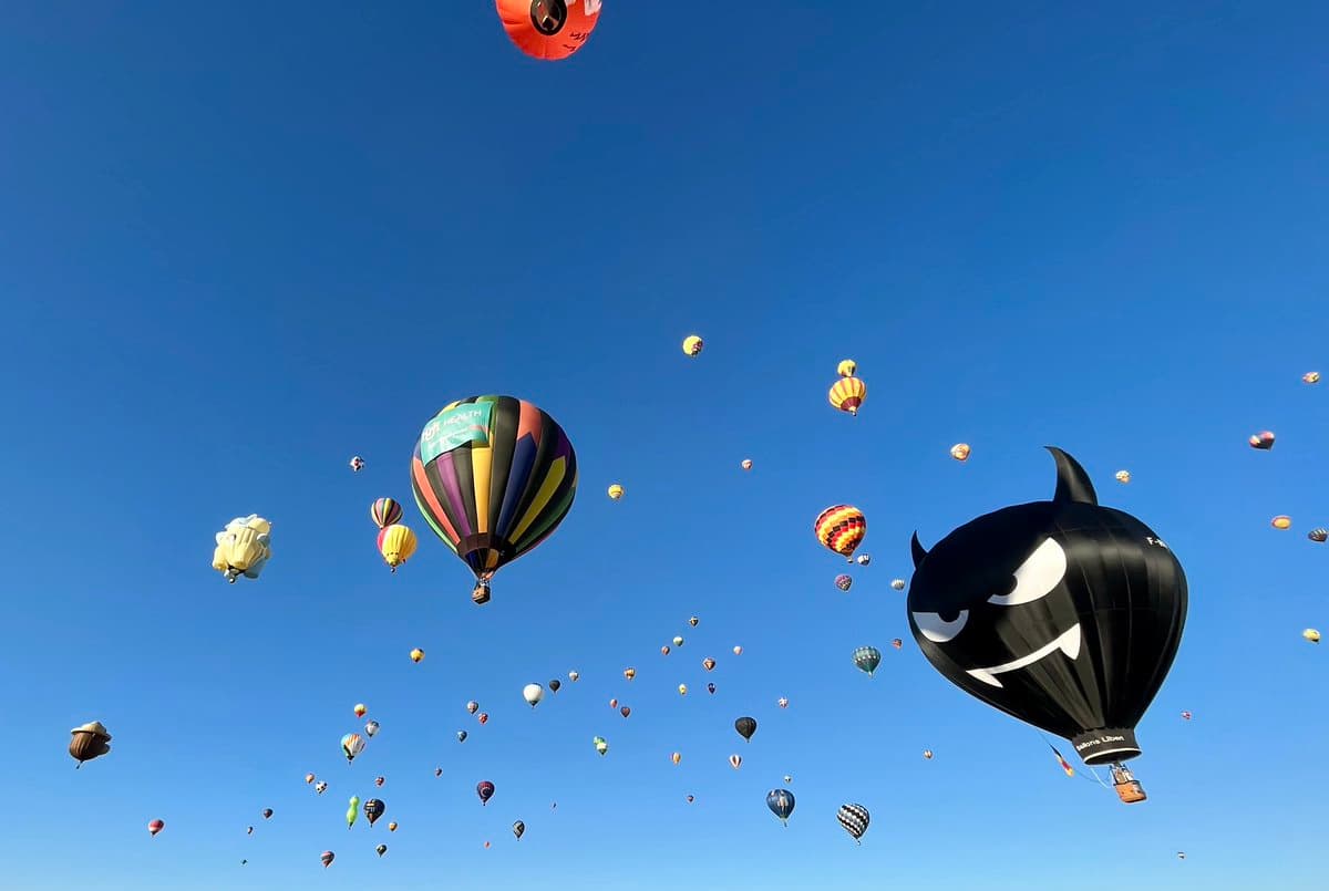 A wide shot of a clear blue sky filled with dozens of hot air balloons at various altitudes. In the foreground, a large black balloon shaped like a devil with horns and a mischievous face floats on the right, while a colorful, striped balloon is visible on the left.