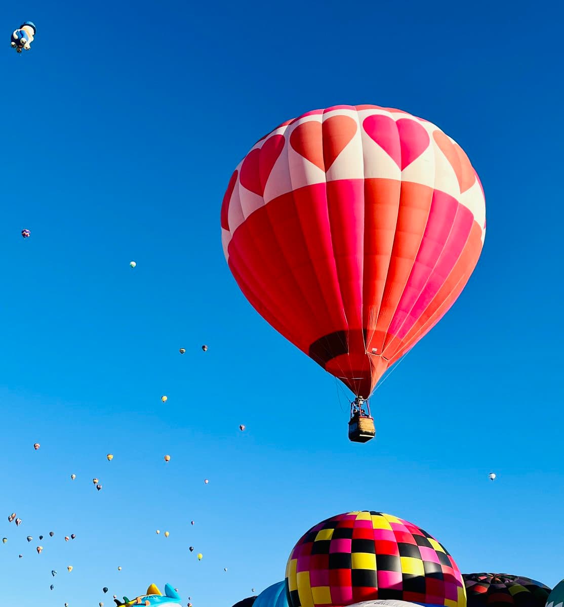 A large hot air balloon decorated with a red and white heart pattern floats in a clear blue sky, surrounded by numerous other balloons in the distance. A colorful, checkered balloon is visible in the foreground at the bottom of the frame.