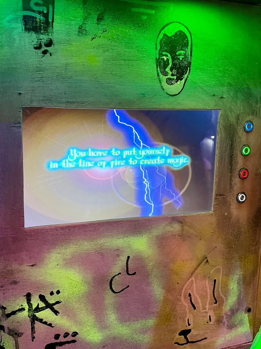 A close-up photograph of a weathered, graffiti-covered arcade cabinet screen displaying a quote. The screen shows a blue lightning bolt graphic and the text 'You have to put yourself in the line of fire to create magic.' against a glowing background.