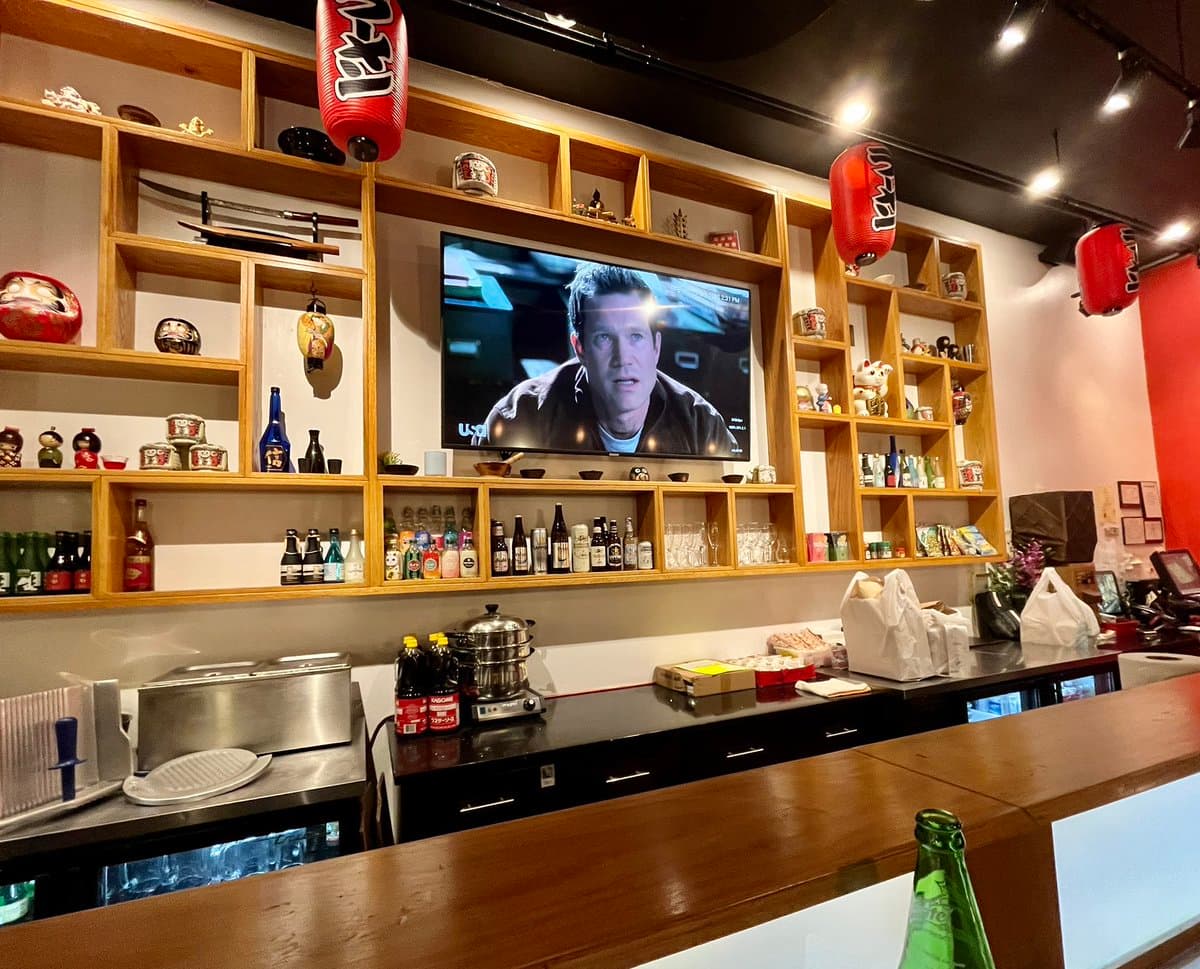 A photo of a Japanese restaurant interior featuring a wooden counter, shelves filled with sake bottles and decorative items, and red paper lanterns. A flat-screen TV mounted on the wall displays a paused scene from a movie or TV show, showing a man in a dark jacket.