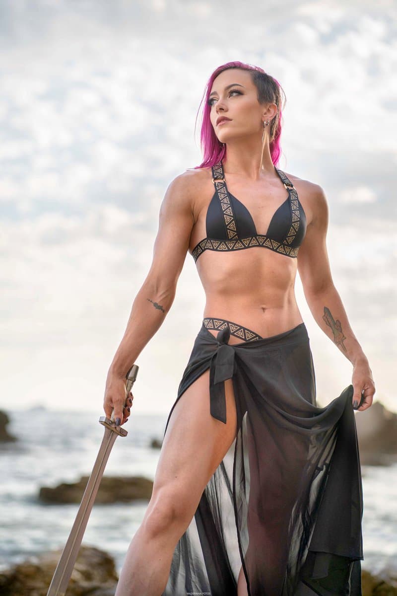 A woman with pink hair stands on a rocky shore holding a sword, wearing a black patterned bikini and a flowing black skirt. The background shows a cloudy sky and the ocean.