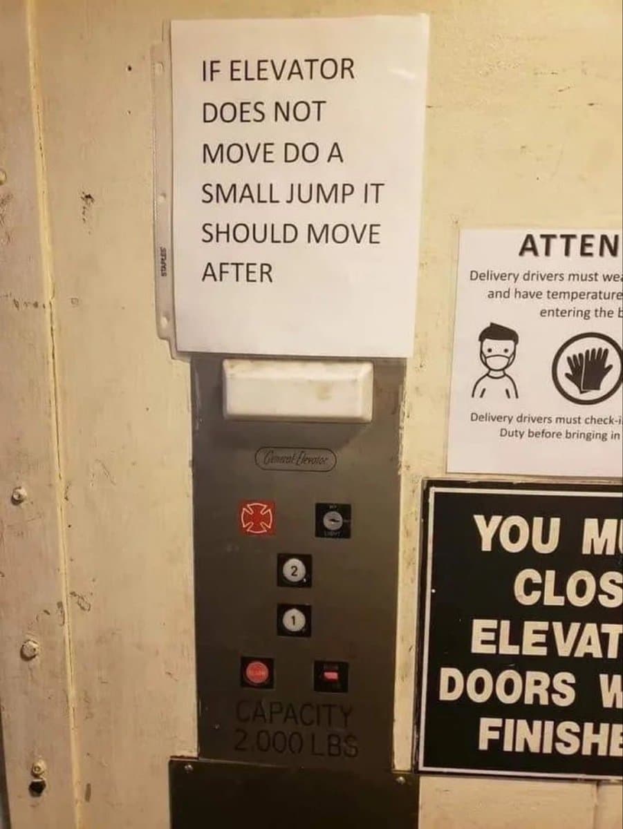 A photograph of an elevator call panel with a handwritten sign taped above it. The sign provides humorous, incorrect instructions for operating the elevator, while other safety and capacity notices are visible on the wall and panel.