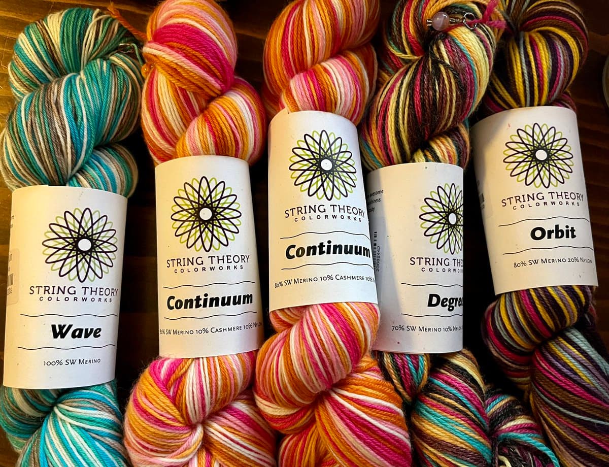 A close-up photograph of five skeins of colorful, variegated yarn from String Theory Colorworks, each with a white paper label. The labels display the brand logo and the names of the yarns: Wave, Continuum, Continuum, Degree, and Orbit.