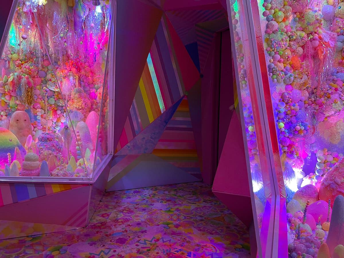 A vibrant, psychedelic interior space featuring walls and floors covered in colorful, textured decorations resembling candy, beads, and glitter. The room is illuminated with bright pink and purple lighting, creating a surreal and whimsical atmosphere.