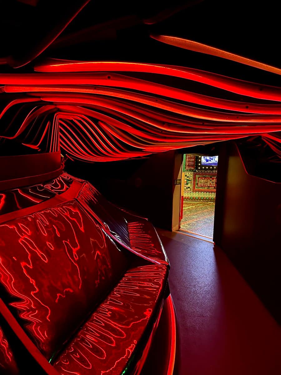 A photograph of a dark interior space featuring a ceiling with wavy, red-lit panels and a glossy, reflective surface in the foreground. An open doorway in the background reveals a brightly lit room with a patterned floor and a wall of illuminated equipment.