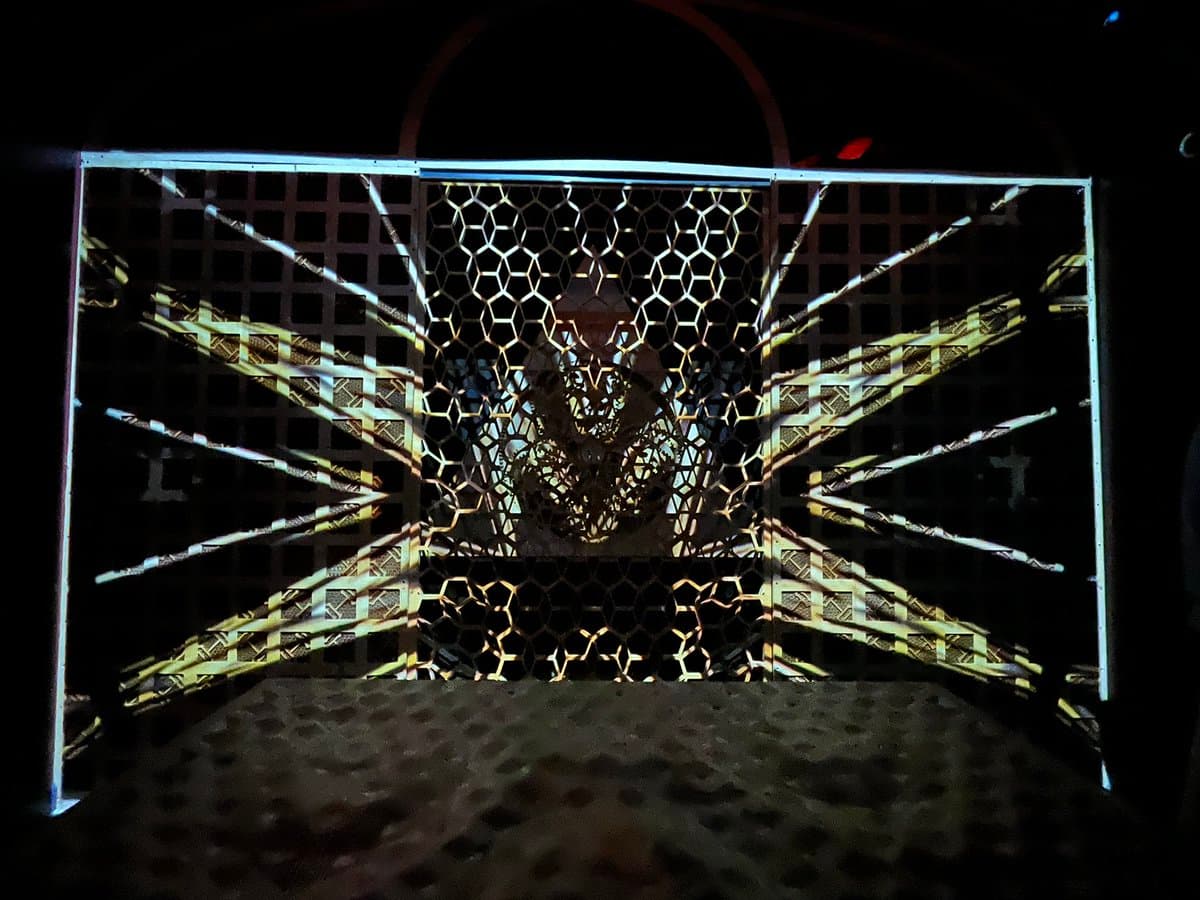 A dark, low-light photograph of a rectangular metal cage or enclosure featuring a grid pattern and a central honeycomb mesh section. The interior is illuminated by a projection of a geometric, starburst-like pattern with radiating lines.
