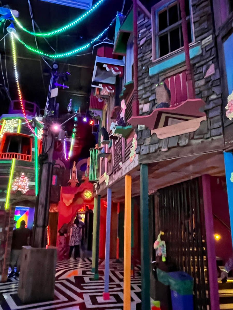 A vibrant, low-angle photograph of a colorful, whimsical indoor attraction featuring stylized buildings, bright neon lights, and a patterned floor.