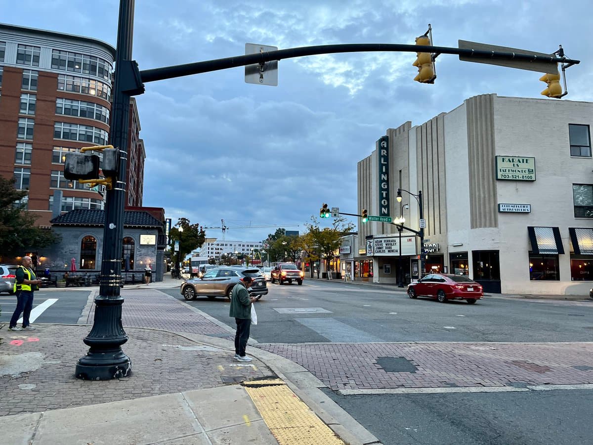 A street-level photograph of an urban intersection on a cloudy day, featuring a crosswalk, traffic lights, and several cars on the road. A person stands on the corner looking at their phone, while a crossing guard is visible on the left. The scene includes a large white building with a vertical sign reading 'ARLINGTON' and a marquee listing movie titles.