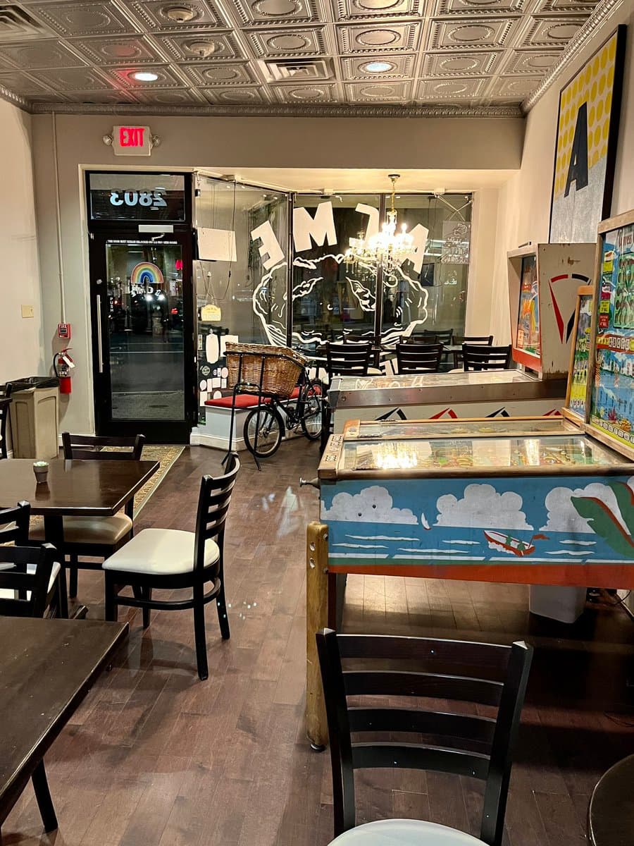 A photo of the interior of a restaurant or cafe, featuring wooden tables and chairs, a decorative tin ceiling, and a glass storefront window with the word "ACME" painted on it. A vintage-style bicycle with a basket is parked near the window, and a pinball machine is visible in the foreground.