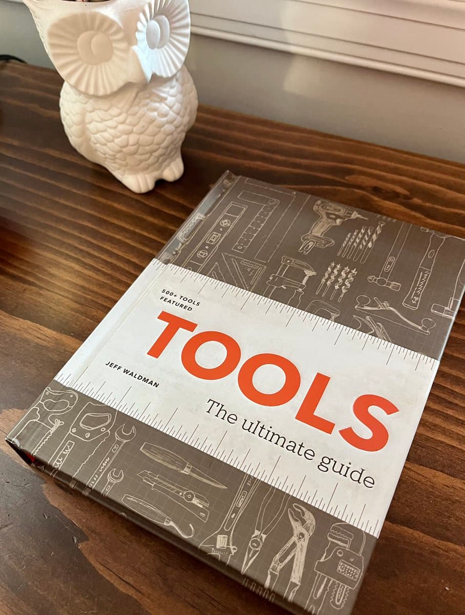 A photograph of a book titled "TOOLS: The ultimate guide" by Jeff Waldman, resting on a wooden table next to a white ceramic owl figurine. The book cover features line drawings of various hand tools and power tools.