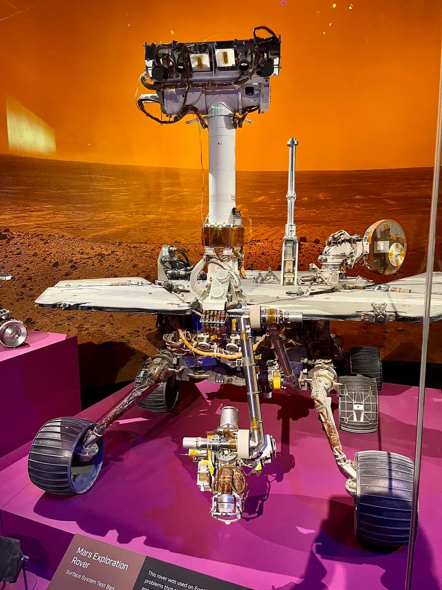 A museum display of a Mars exploration rover on a purple platform, set against a backdrop of a reddish-orange Martian landscape. A small informational placard is visible in the lower left corner.
