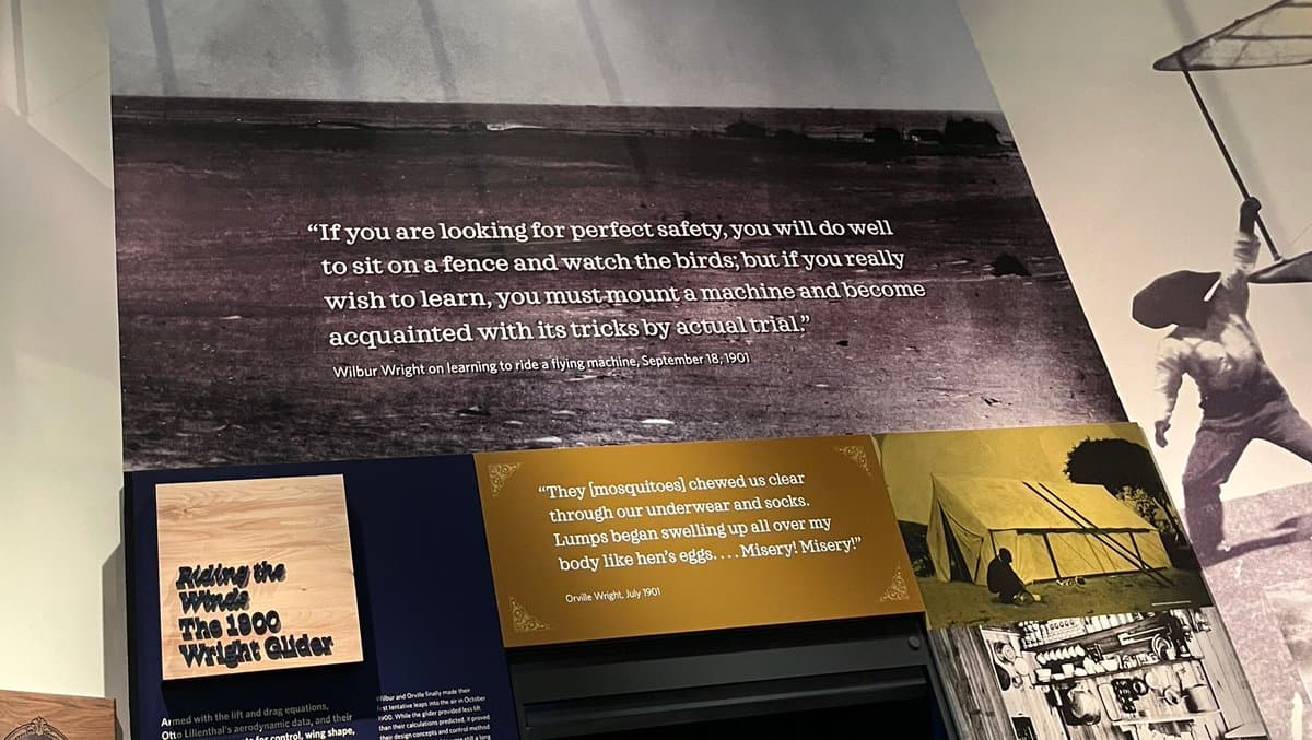 A museum display panel featuring historical black-and-white photographs of early aviation and text panels with quotes from the Wright brothers. The main panel shows a quote from Wilbur Wright about learning to fly, while a lower section includes a sign titled "Riding the Winds: The 1900 Wright Glider" and another quote from Orville Wright about mosquitoes.