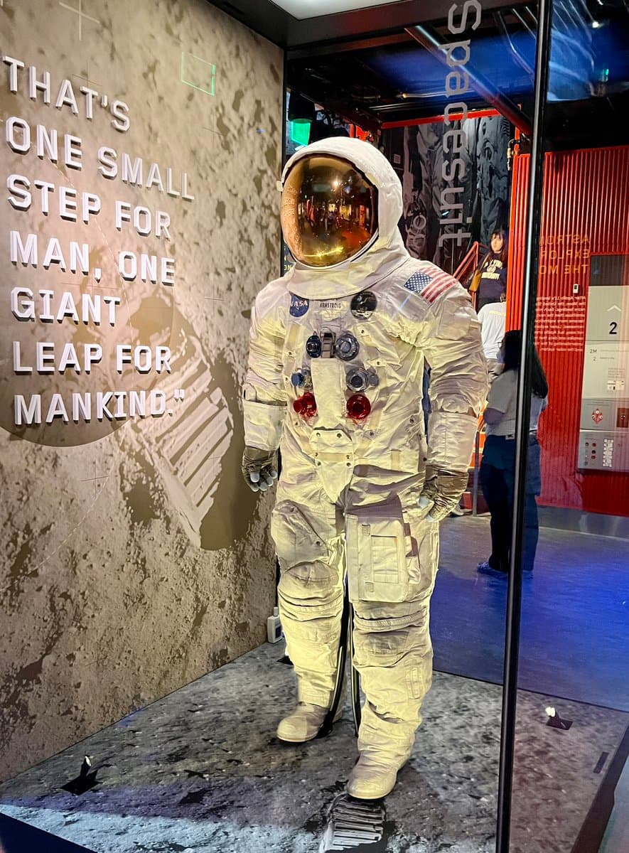 A photograph of a museum exhibit featuring a full astronaut spacesuit standing on a lunar surface display. The suit is positioned in front of a wall with a large quote and a backdrop showing a space capsule.