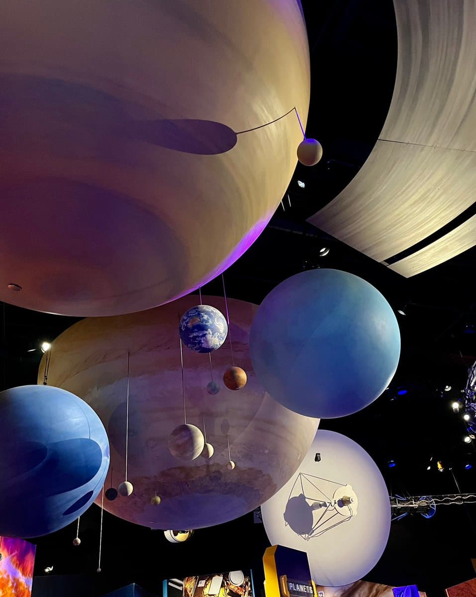 A low-angle photograph of a museum exhibit featuring large, suspended spheres representing planets and moons against a dark background. The scene includes a prominent model of Earth and a large, textured sphere resembling Jupiter or Saturn, with smaller planets hanging on wires. A sign at the bottom right reads "PLANETS".