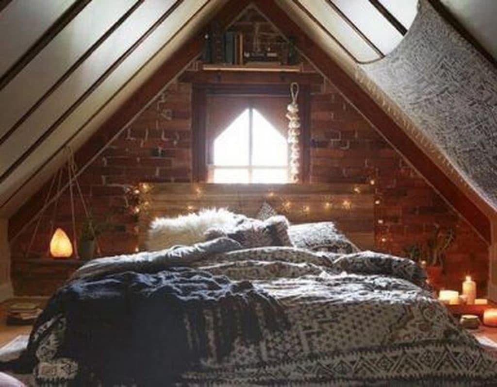 A cozy attic bedroom featuring a bed with patterned blankets, exposed brick walls, and a small window, illuminated by string lights and candles.