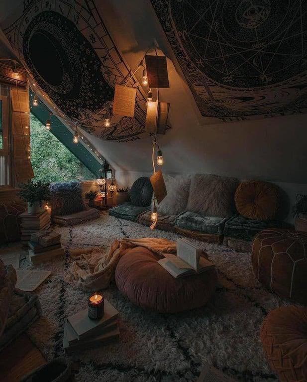 A cozy, dimly lit attic room decorated with bohemian-style elements, including a large tapestry on the slanted ceiling, string lights, floor cushions, and a plush rug. An open book rests on a large floor cushion in the center, surrounded by stacks of books and a lit candle.