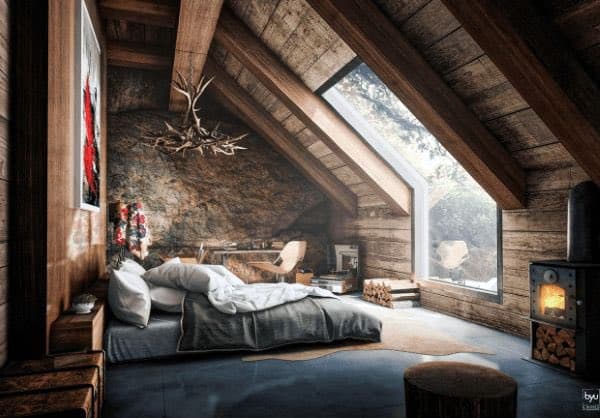A cozy, rustic bedroom interior featuring a low bed with white bedding, a wooden ceiling with exposed beams, and a large window looking out onto a snowy landscape. A wood-burning stove with a visible fire sits in the corner, and a chandelier made of antlers hangs from the ceiling.