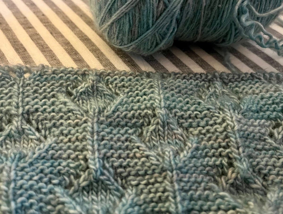 A close-up photograph of a partially completed knitting project featuring a textured pattern in shades of blue and grey, with a ball of matching yarn resting on a striped cloth in the background.