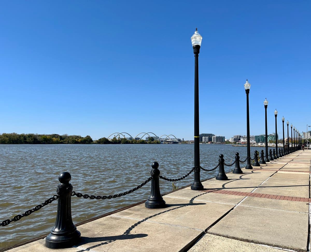 A scenic outdoor photograph of a paved waterfront promenade on a sunny day, featuring a row of black lampposts and chain-link bollards along the edge of a river with a city skyline and bridges in the distance.