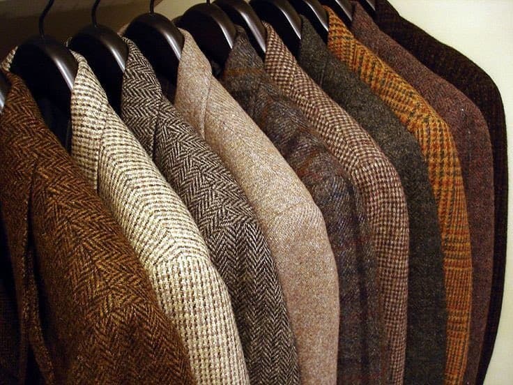 A row of men's wool coats in various earth tones and patterns, including herringbone and plaid, hanging on black hangers.