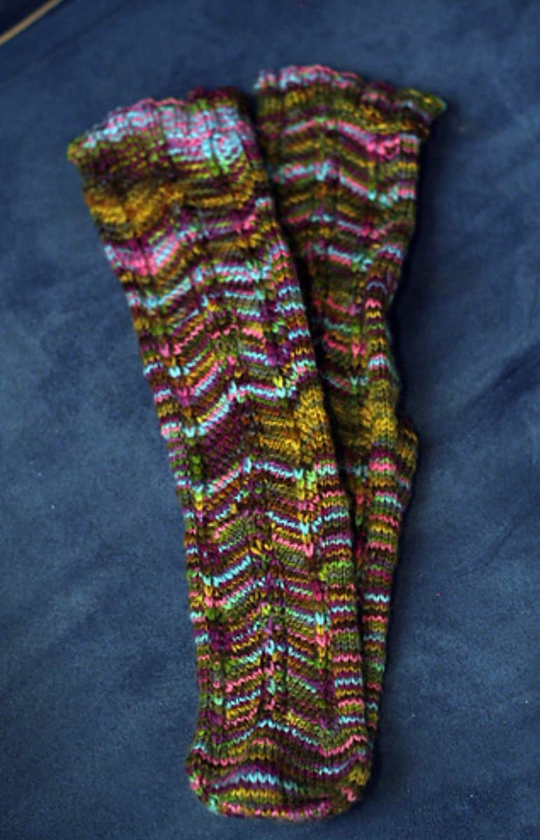A pair of hand-knitted socks featuring a colorful, multi-toned chevron pattern in shades of pink, purple, green, and yellow, laid flat on a dark blue fabric surface.