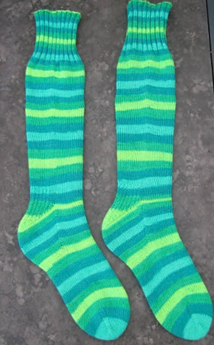 A pair of hand-knitted socks with horizontal stripes in shades of green and blue, laid flat on a gray concrete surface. The socks feature ribbed cuffs and heels, showcasing a simple yet colorful striped pattern.
