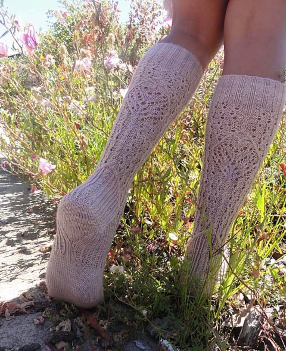 A close-up photograph of a person's lower legs and feet wearing a pair of cream-colored, lace-knit socks. The socks feature an intricate, openwork pattern and are shown outdoors against a background of green foliage and pink flowers.