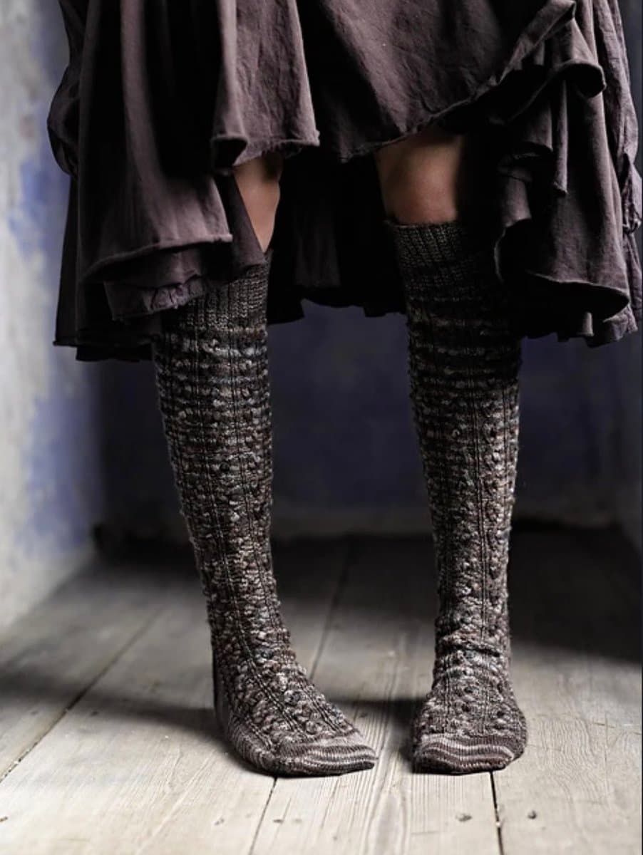 A person stands on a wooden floor wearing a dark, layered skirt and tall, intricately knitted socks that reach up to the knees. The socks feature a textured, patterned design in shades of gray and black.