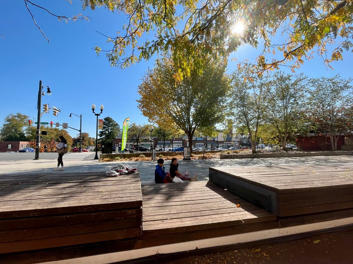 A sunny outdoor photograph of a public plaza featuring large wooden benches, with a few people sitting and walking around. In the background, there is a street intersection with traffic lights, cars, and storefronts lined with trees showing early autumn foliage.