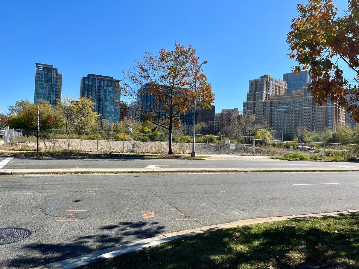 A photograph of a fenced-off, empty lot with dry grass and a single tree, situated across a street from a row of modern high-rise apartment buildings under a clear blue sky.