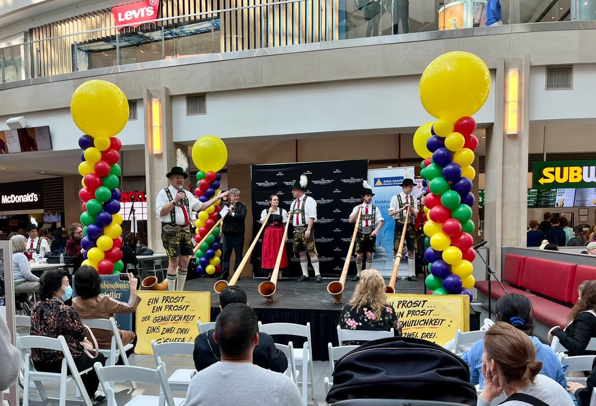 A live performance by a traditional German brass band on a stage in a shopping mall atrium, decorated with tall columns of colorful balloons. The musicians, dressed in traditional dirndls and lederhosen, are playing long alphorns in front of a seated audience.