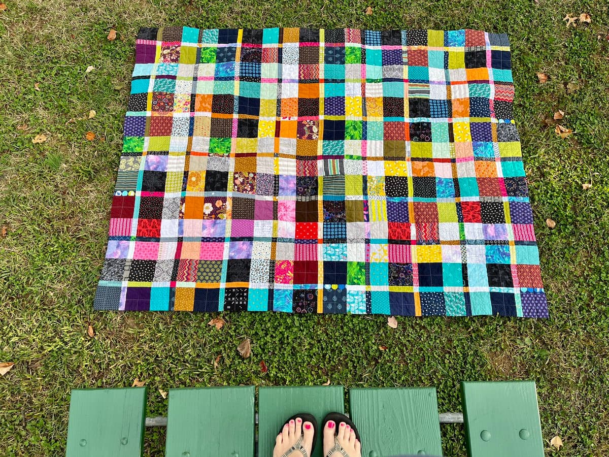 A top-down view of a colorful, patchwork quilt laid out on a grassy lawn. The bottom of the frame shows a person's feet in sandals resting on a green park bench.